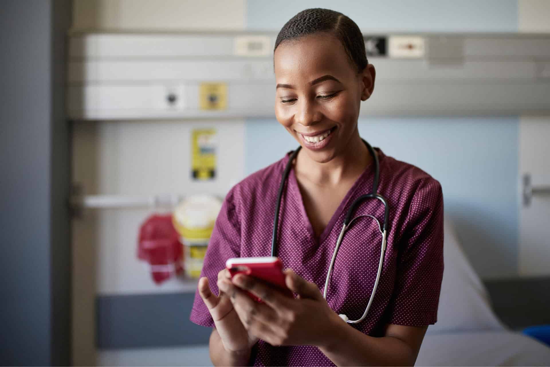 Young Black nurse in purple scrubs wearing stethoscope scrolling on smartphone