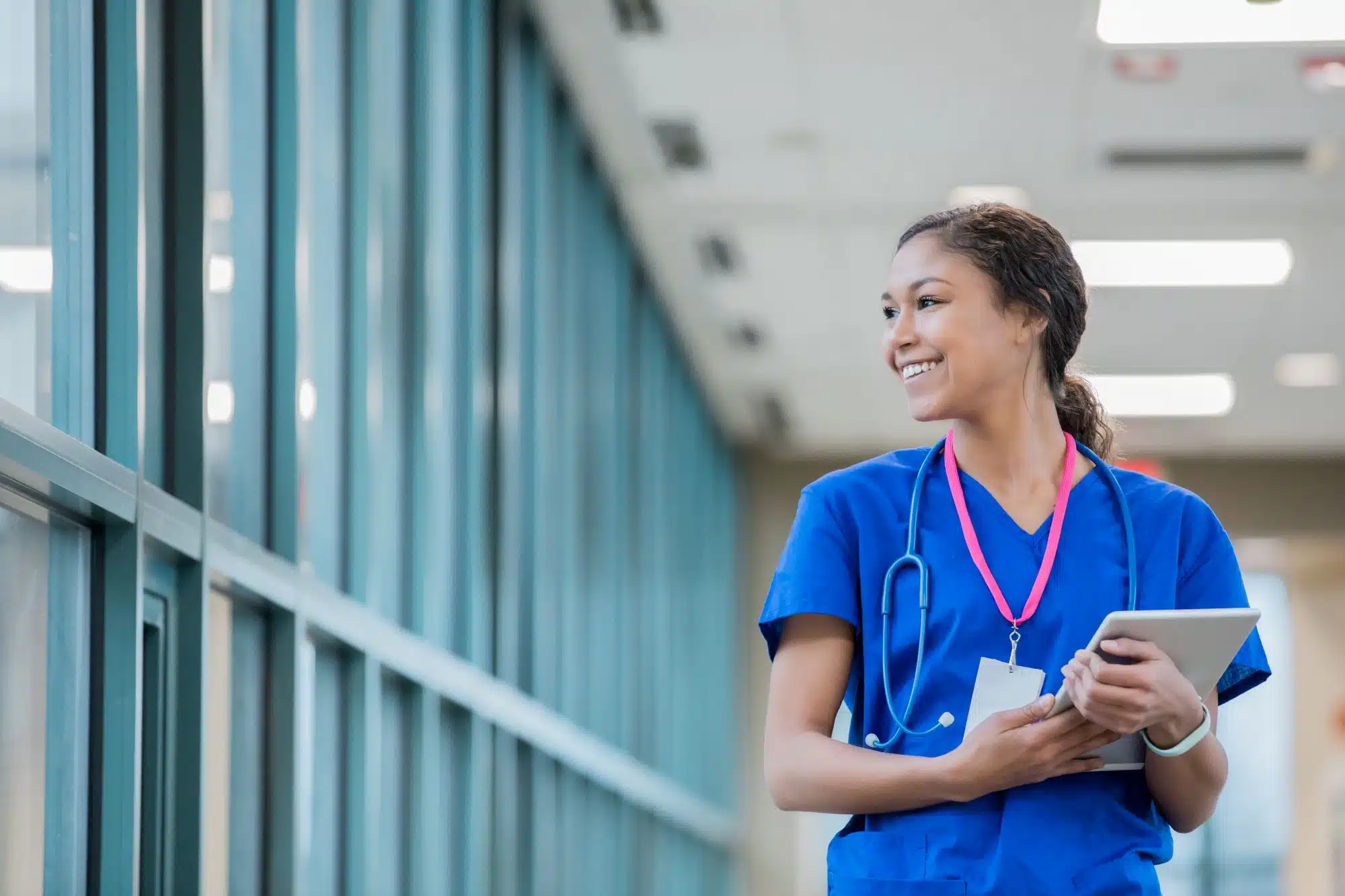Young nurse wearing stethoscope, carrying tablet, walking through hospital hallway
