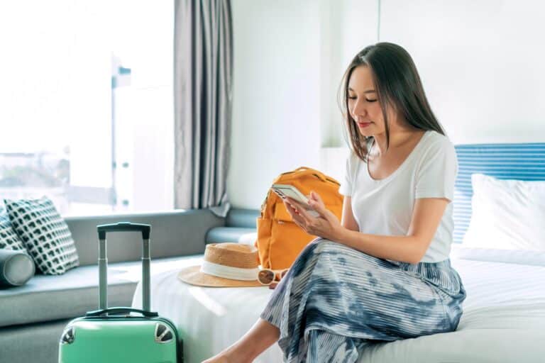 Happy young Asian traveler woman using mobile phone while relaxing on bed in hotel room next to suitcase and backpack