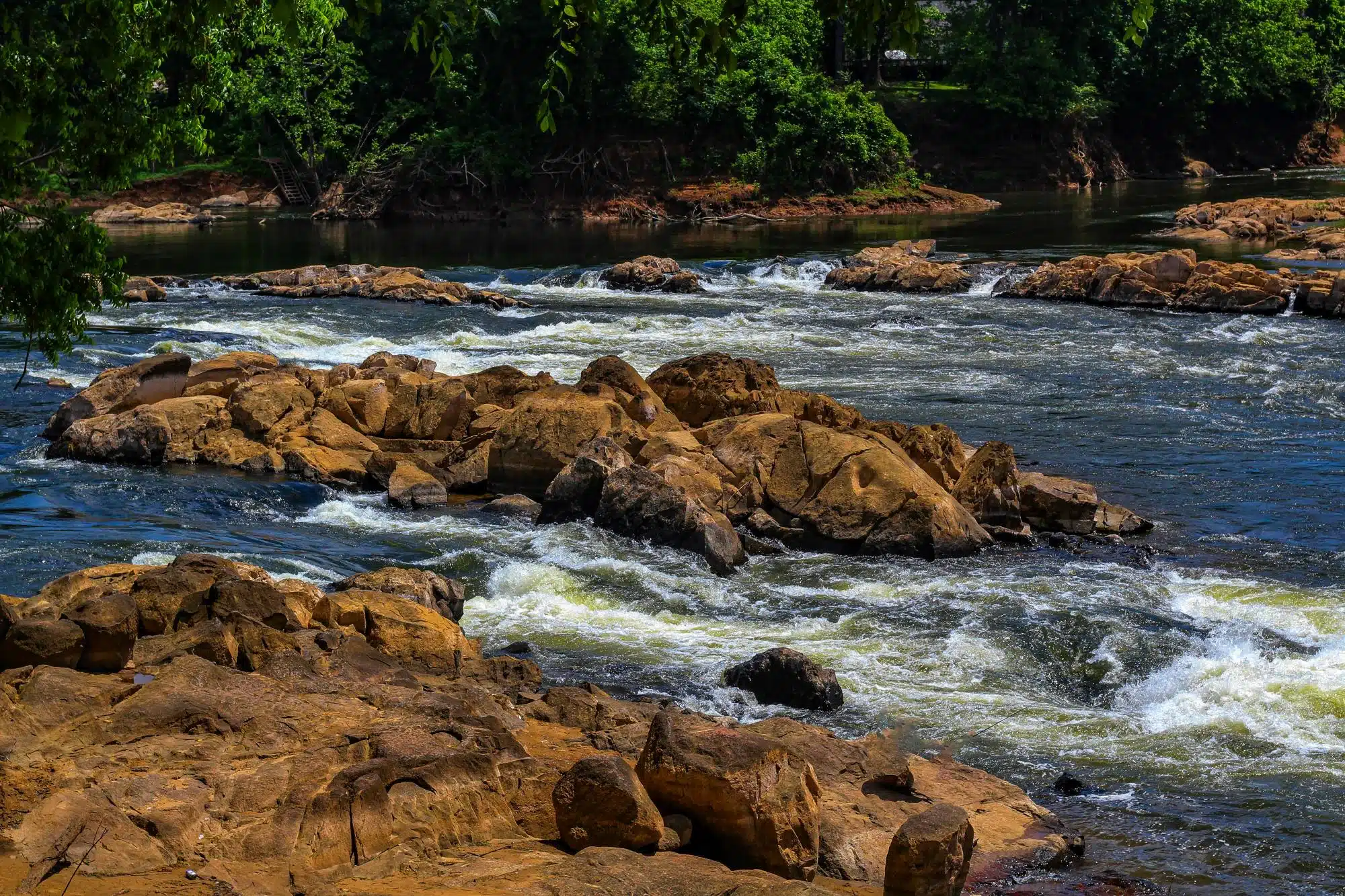 Large boulders in the river with flowing water and greenery in the background on a bright sunny day