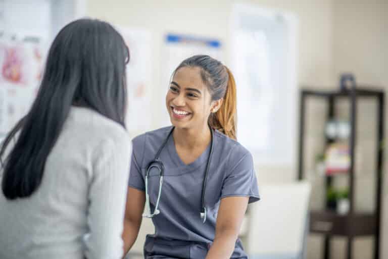 A young female nurse of Asian descent is smiling widely at her patient who is sitting across her. She is wearing medical scrubs and has a stethoscope around her neck.