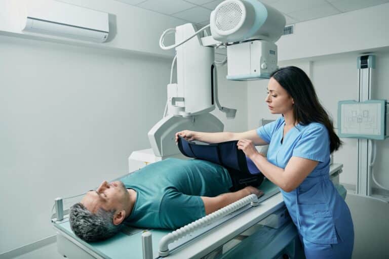 Female technician preparing patient for body X-ray in radiographic imaging room, putting for him radiation protection apron