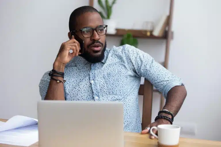 Young Black businessman talking on phone in front of laptop at desk.